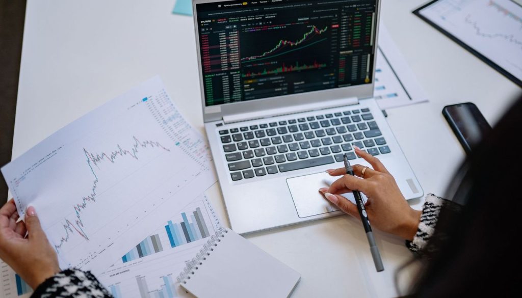 Un ordinateur portable affichant des courbes de cours de Bourse, posé sur un bureau à côté d'un carnet de notes et d'un stylo.