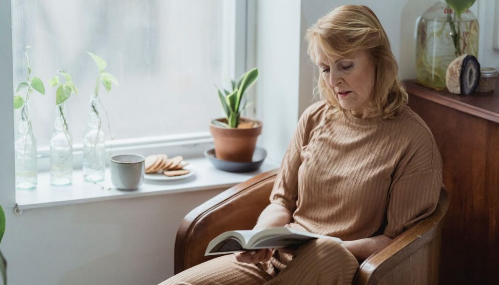 Une femme lit un livre dans un fauteuil près d'une fenêtre, une tasse posée à côté d'elle